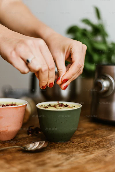 Hands sprinkle spices over yellow soup.